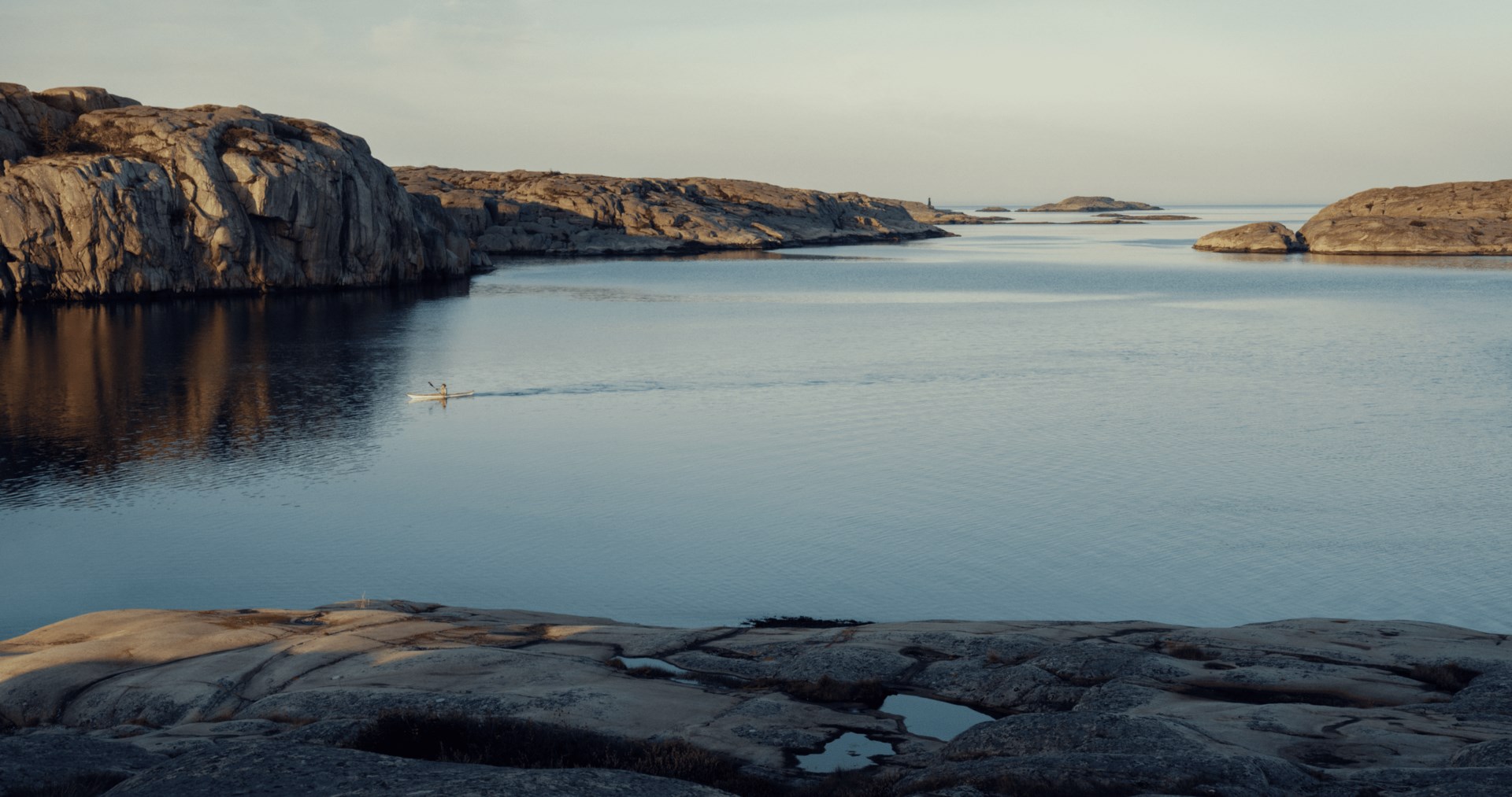 Paysage côtier paisible avec formations rocheuses lisses et un kayakiste solitaire pagayant sur une eau calme au coucher du soleil.