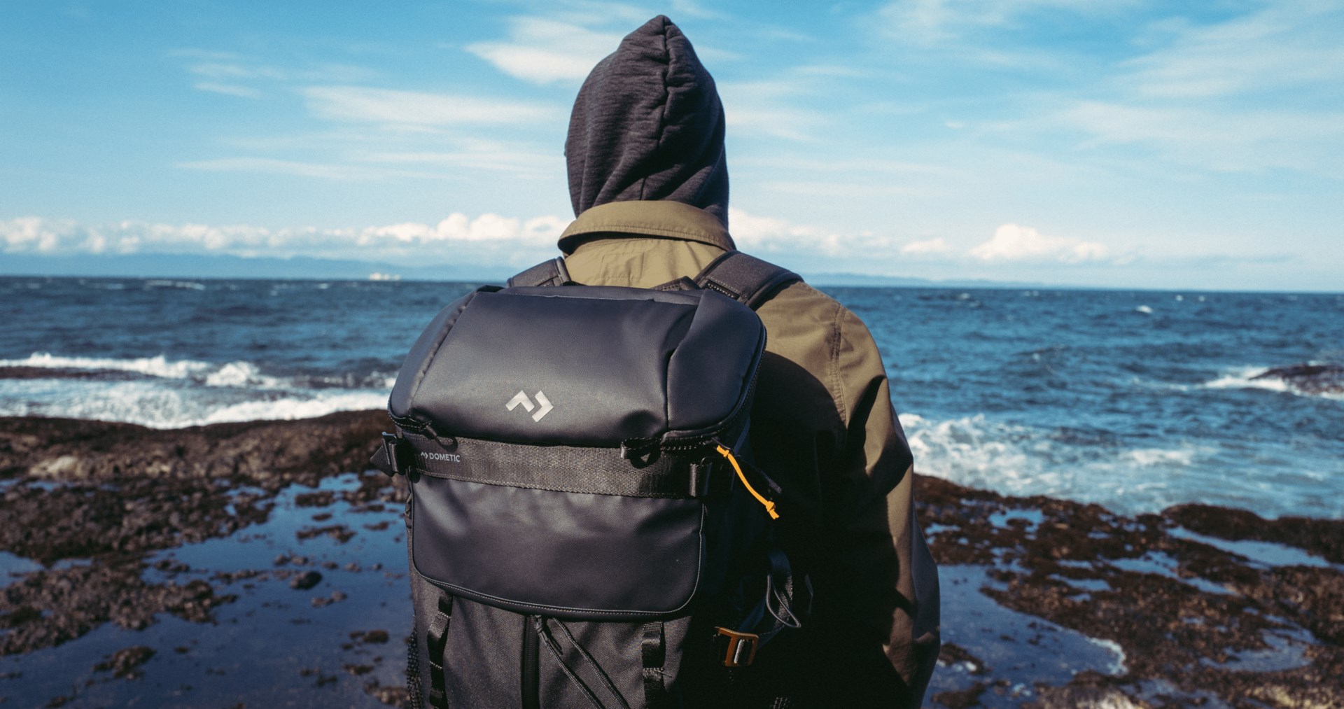 Personne portant une veste à capuche et un sac isotherme Dometic Unrestricted sur le dos, au bord d’une côte rocheuse